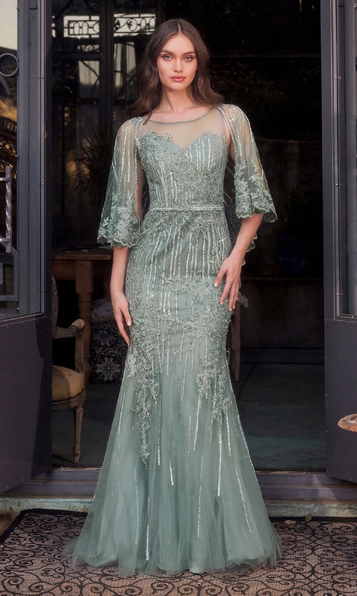 A woman stands in a doorway wearing the Andrea & Leo MA105 Cape-Sleeve Beaded Formal Gown, a floor-length sage green dress featuring intricate beadwork and sheer cape sleeves.